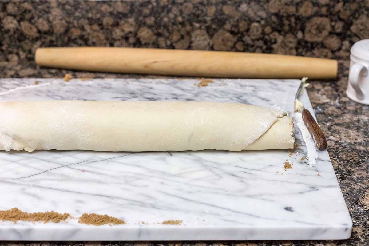 Homemade cinnamon rolls rolled into a long tight spiral and placed on a marble board. A rolling pin is behind the board.