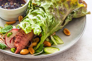 Low carb lettuce wrap with sliced steak, mint, radish, cucumber and peanuts wrapped up on a plate. A dipping sauce is on the plate as well.