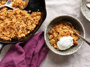 Bowl of 3-ingredient skillet apple crisp with a dollop of yogurt next to a cast iron skillet with more apple crisp, a purple kitchen towel, and a bowl