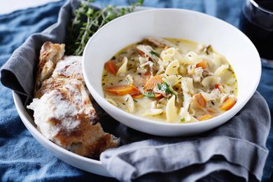 A white bowl filled with the best creamy chicken noodle soup set on a grey linen against a blue background with a bundle of thyme near by and a hunk of sourdough bread..