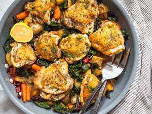 Close up photo of of crispy chicken thighs with cubed potatoes and greens visible in the dish. Serving fork and spoon are on the upper edge of the skillet. The chicken is sprinkled with herbs and a striped grey linen is behind the skillet.