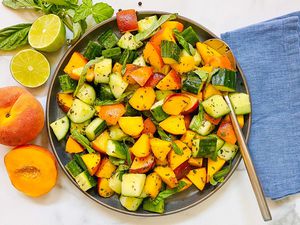 Summer peach and cucumber salad in a bowl with a serving spoon. A blue linen is to the right and peaches, limes and basil are to the left of the bowl.