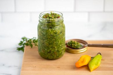 Jar of Carribbean green seasoning next to some peppers and a jar lid with a spoon resting on it, all on a cutting board