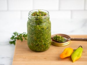 Jar of Carribbean green seasoning next to some peppers and a jar lid with a spoon resting on it, all on a cutting board