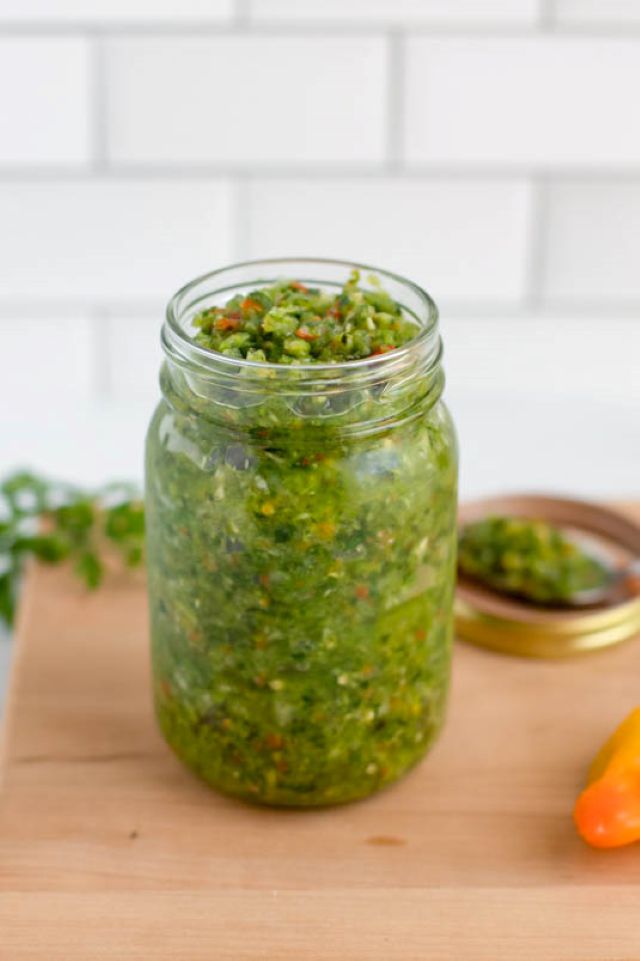 Jar of Carribbean green sauce surrounded by peppers, a jar lid, and parsley, all on a cutting board