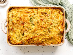 Potato Kugel in a Casserole Dish Surrounded by a Small Saucer with Herbs, Another Small Saucer with Cracked Pepper, and a Kitchen Linen