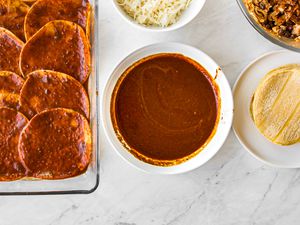 Bowl of Red Enchilada Sauce Surrounded by Enchilada Ingredients (a Plate of Tortillas, Bowl of Shredded Chicken, Bowl of Shredded Cheese, and Glass Platter with Sauce Soaked Tortillas)