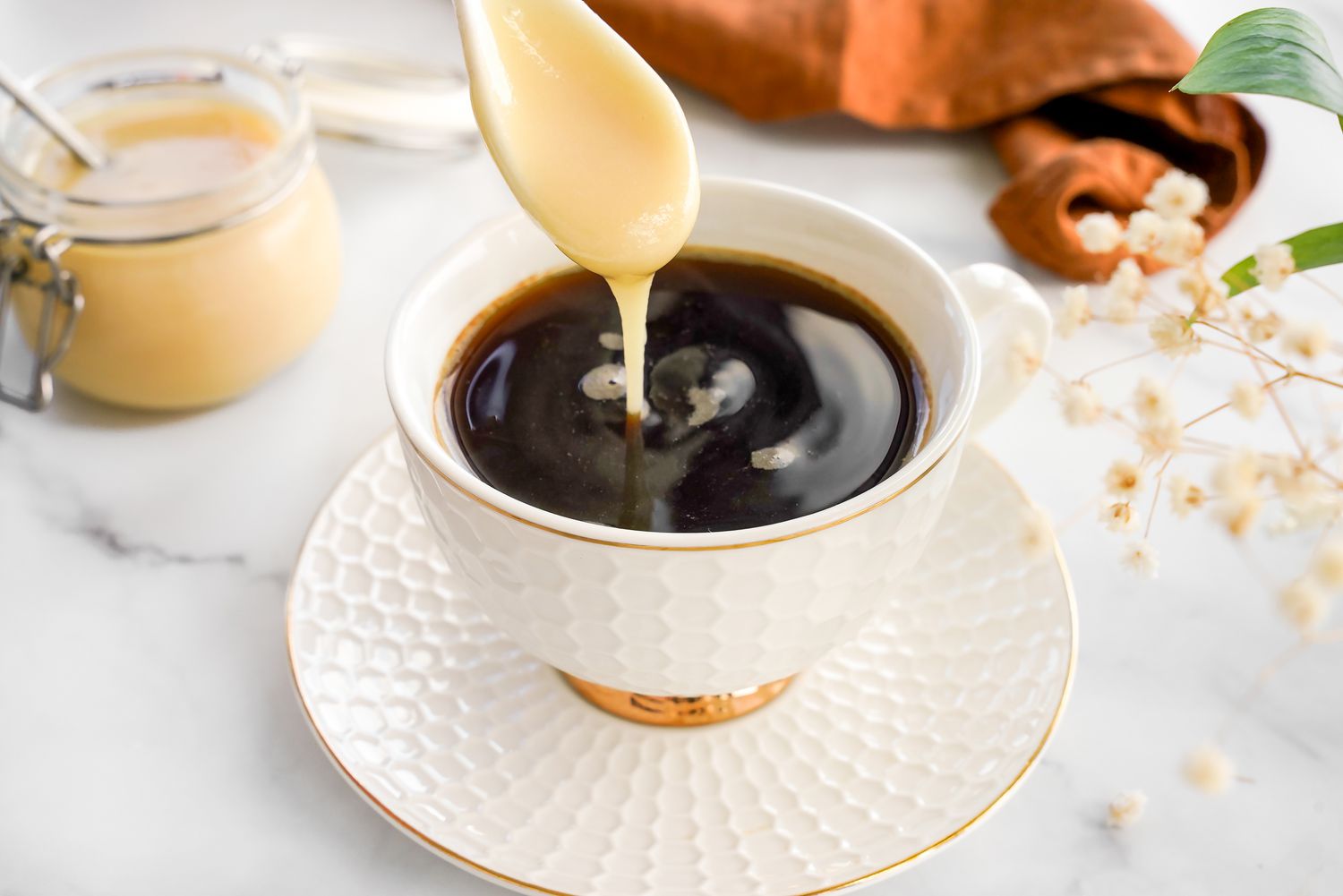 Spoonful of Homemade Condensed Milk Poured Into a Cup of Coffee, and in the Background, a Kitchen Towel and a Jar With More Condensed Milk