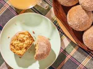 A halved apple cider donut muffin on a plate, and in the surroundings, a cup of apple cider and more muffins stacked on a plate