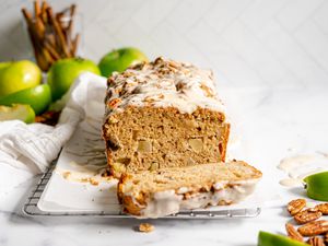 Apple Fritter Bread with a Slice Cut Off on Parchment Paper Surrounded by Granny Smith Apples, Pecans, and Sticks of Cinnamon