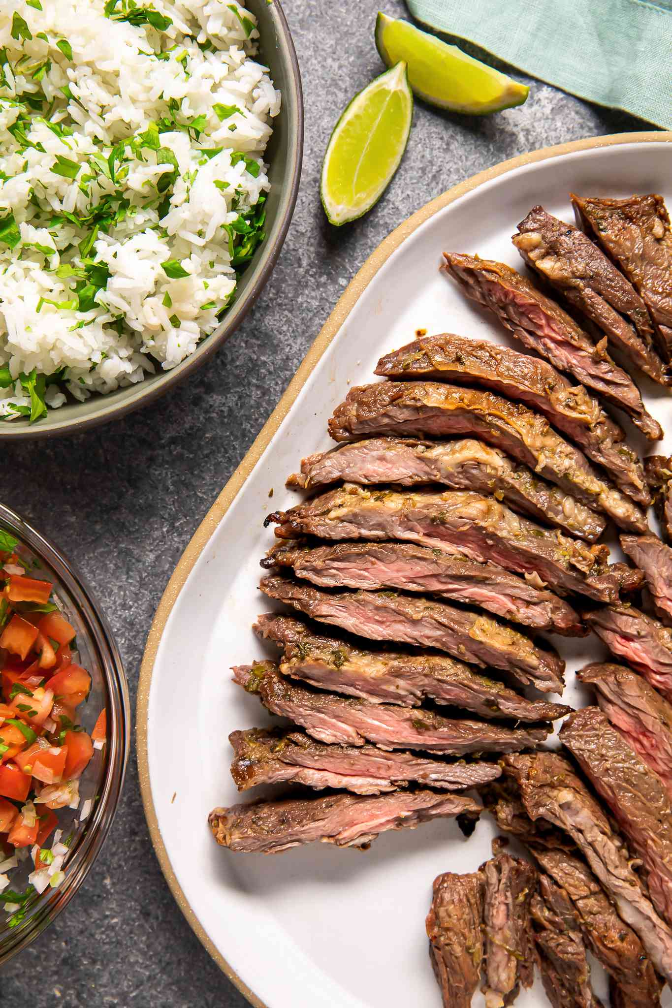 Arrachera (Mexican Skirt Steak) on a Platter Next to a Bowl of Salsa, a Bowl of Cilantro Rice, and Lime Wedges on the Counter