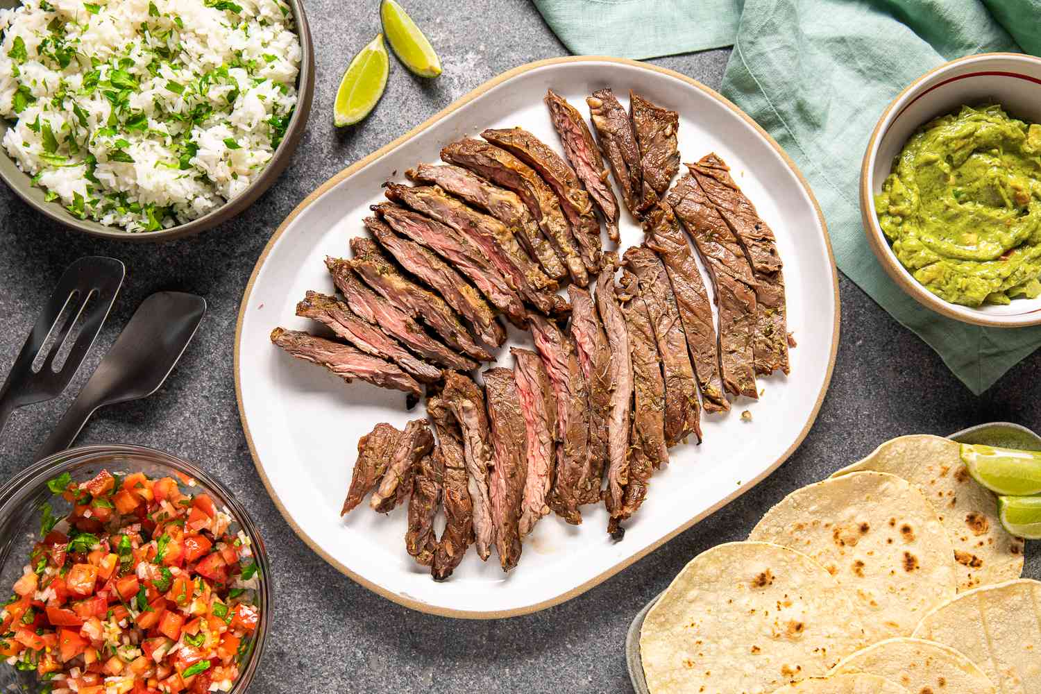 Arrachera (Mexican Skirt Steak) on a Platter Next to a Bowl of Salsa, a Bowl of Cilantro Rice, a Bowl of Guacamole, a Platter of Tortillas, and Lime Wedges and a Light Teal Kitchen Towel on the Counter