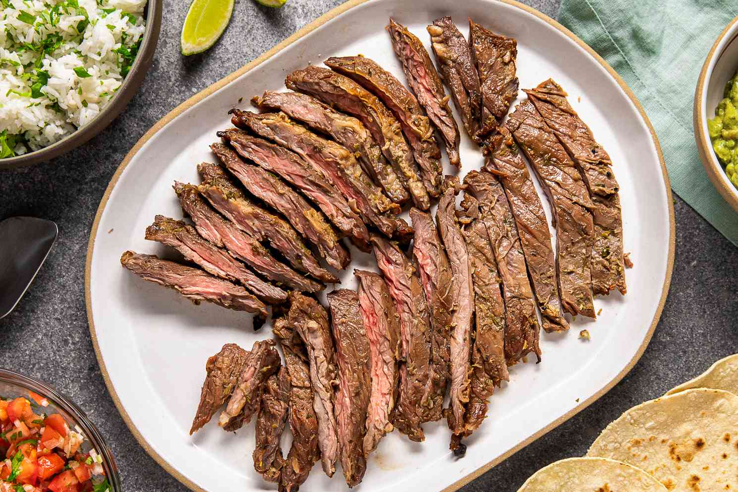 Arrachera (Mexican Skirt Steak) on a Platter Next to a Bowl of Salsa, a Bowl of Cilantro Rice, a Bowl of Guacamole, a Platter of Tortillas, and Lime Wedges and a Light Teal Kitchen Towel on the Counter