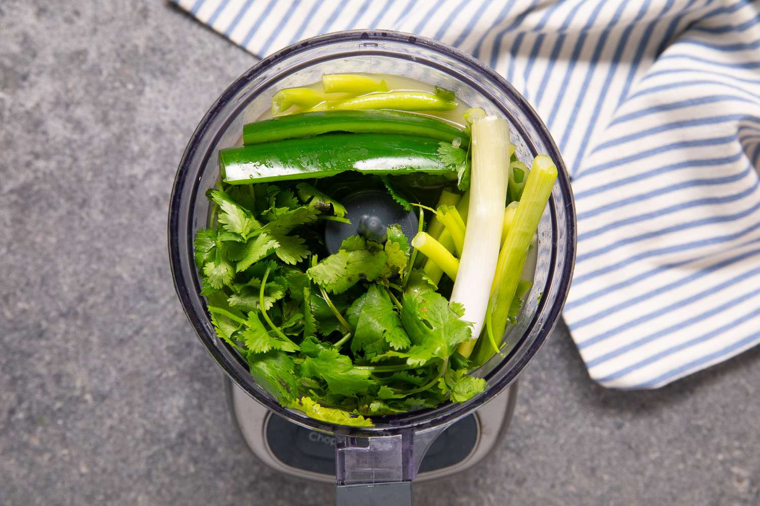 Cilantro, Green Onions, and Serrano Peppers in a Food Processor Next to a Blue and White Stripped Kitchen Towel 