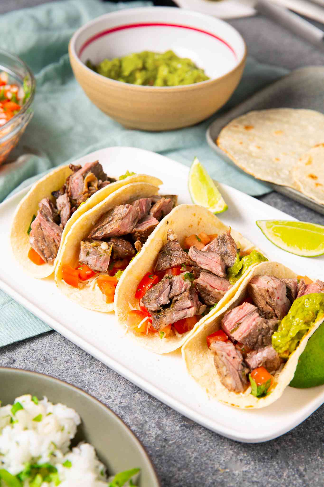 Platter of Arrachera (Mexican Skirt Steak) Tacos With Lime Wedges, and in the Surroundings, a Bowl of Cilantro Rice, a Bowl of Salsa, a Bowl of Guacamole, and a Tray of Tortillas, All on a Light Teal Kitchen Towel