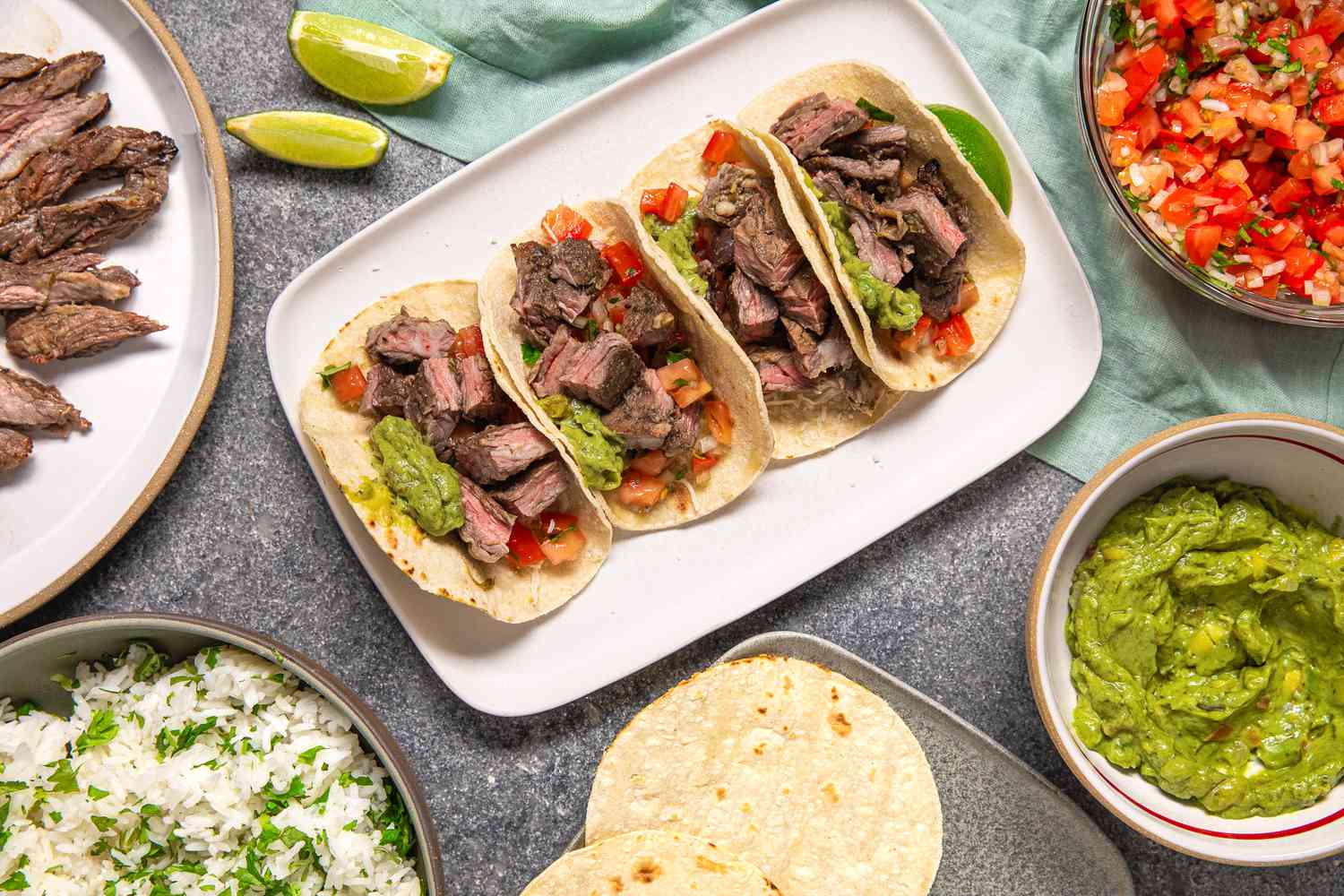 Platter of Arrachera (Mexican Skirt Steak) Tacos With Lime Wedges, and in the Surroundings, a Platter of Arrachera (Mexican Skirt Steak), a Bowl of Cilantro Rice, a Bowl of Salsa, a Bowl of Guacamole, and a Tray of Tortillas, All on a Light Teal Kitchen Towel