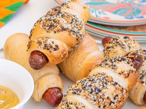 Homemade Hot Dog Bagels (Some With Everything Bagel Topping and Some Plain) on a Plate, and in the Surroundings, a Stack of Plates and a Bowl of Yellow Mustard 