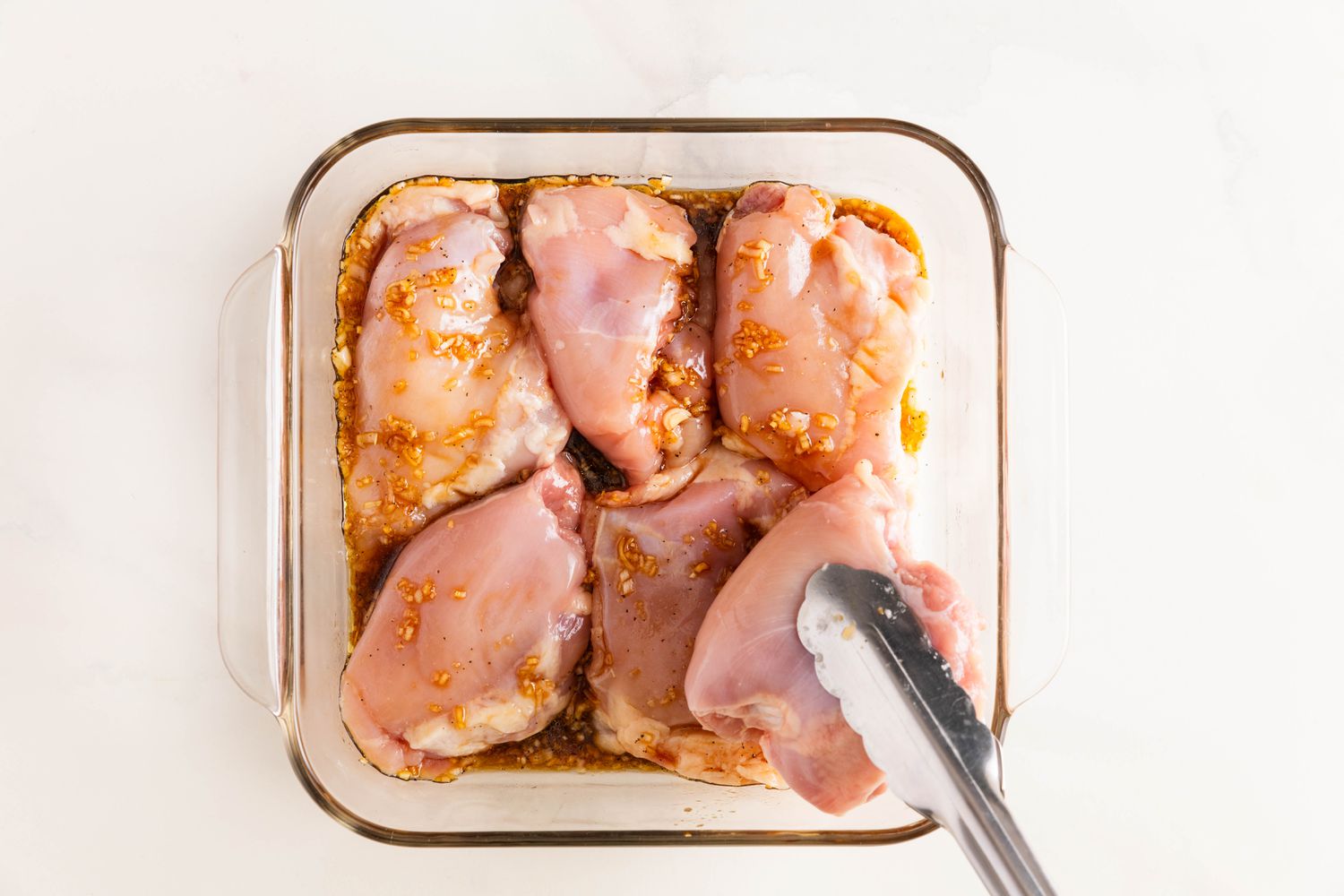 Tongs adding chicken thighs to a glass Pyrex dish with marinade for banh mi salad recipe