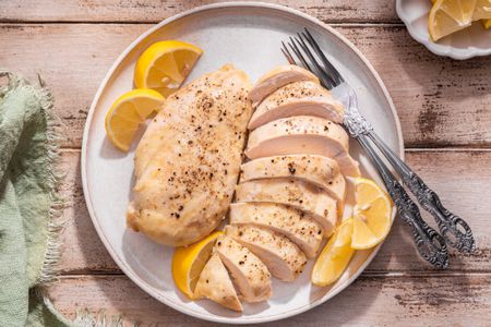 Chicken breasts (one cut into slices) on a plate with lemon wedges and utensils, and in the surroundings, a sage kitchen linen and a small saucer with more lemon wedges