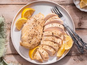 Chicken breasts (one cut into slices) on a plate with lemon wedges and utensils, and in the surroundings, a sage kitchen linen and a small saucer with more lemon wedges