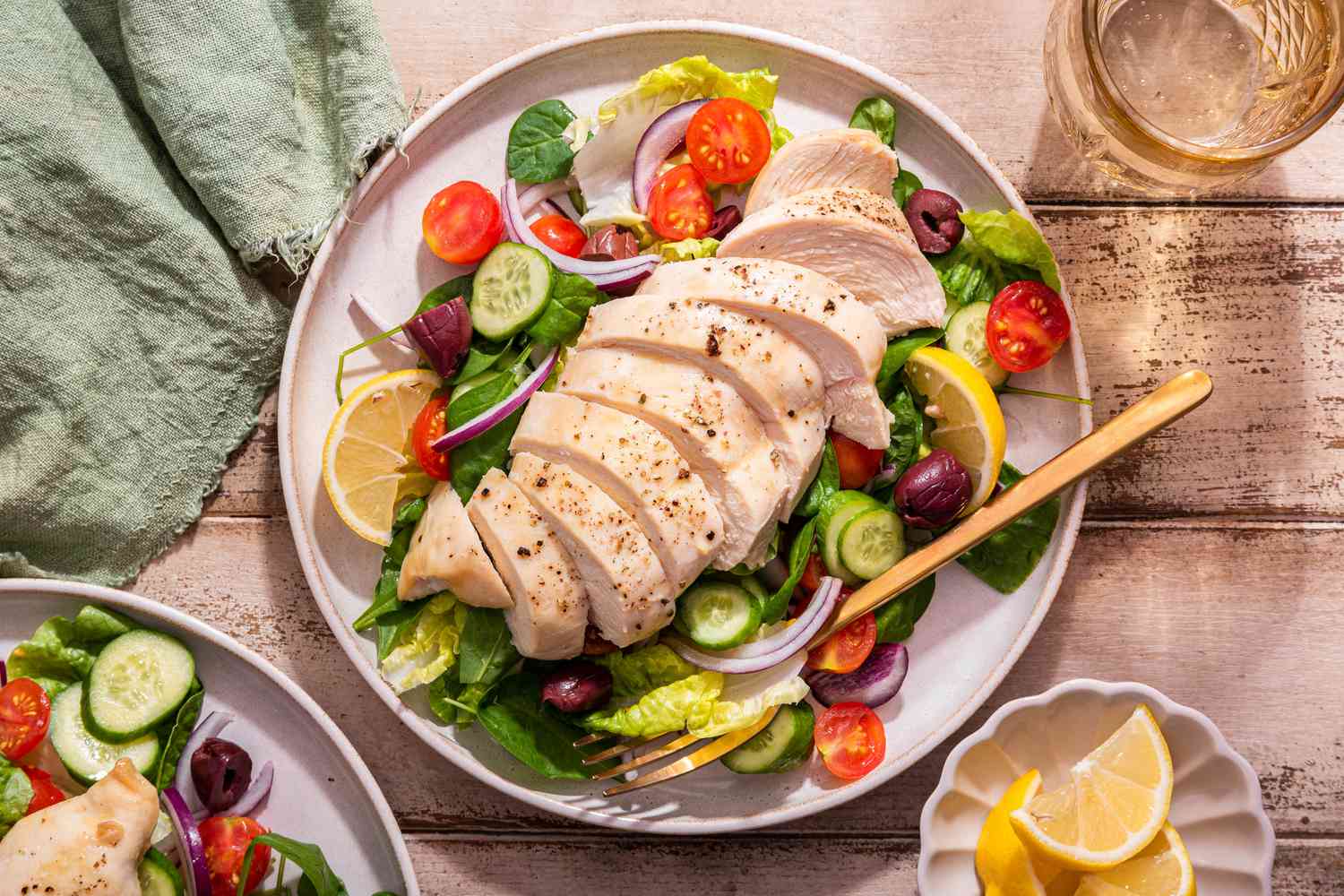 Sliced baked chicken breast on a plate with a salad, and in the surroundings, a small saucer with lemon slices, another serving of chicken and salad on a plate, a sage kitchen towel, and a glass of water