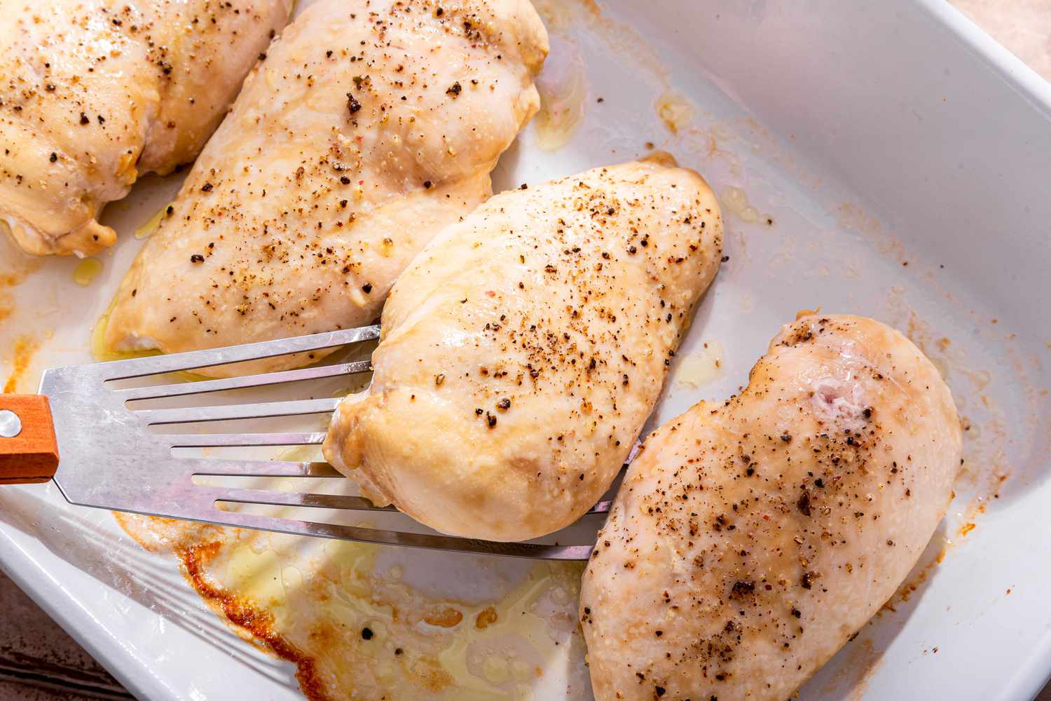 Close-up: baked chicken breast in a casserole dish with one piece slightly lifted using a spatula 