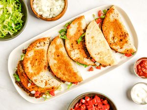 Baked tacos on a platte surrounded by bowls of toppings (bowl of shredded iceberg lettuce, bowl of diced onions, bowl of tomatoes, bowl of salsa, and a bowl of sour cream)