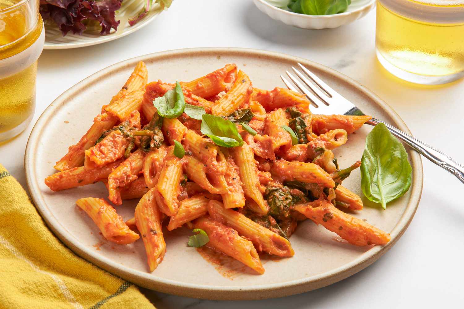 One pan baked penne alla vodka on a plate at a table setting with a plate of side salad, two drinks, and a small plate of basil