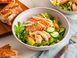 Banh mi salad and in the surroundings, another bowl, a wooden board with baguette slices, and a table napkin with a fork