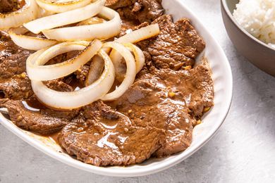 Plate of Bistek Topped With Onions Next to a Bowl of Rice