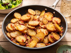 Panful of Bratkartoffeln (German Cottage Fries) at a rustic table setting with a bowl of salad, a glass of water, and a small saucer with salt and pepper