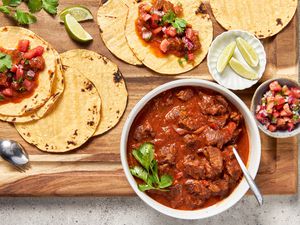 On a wooden board, a bowl of carne picada with a sprig of cilantro and a spoon, surrounded by a bowl of pico de gallo, a small plate of lime wedges, and tortillas spread all over the board (some with carne picada and pico de gallo, some plain)