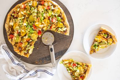 Slice of Cheeseburger Pizza on Small Plate Next to a Circular Wooden Tray with More Pizza and a Pizza Wheel and on the Counter, a Kitchen Towel