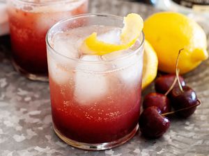 Homemade cherry pop cocktail with fresh cherries and lemon set next to it.