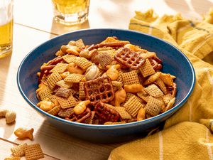Sweet and spicy chex mix in a bowl at a table setting with two glasses of beer and a yellow kitchen towel