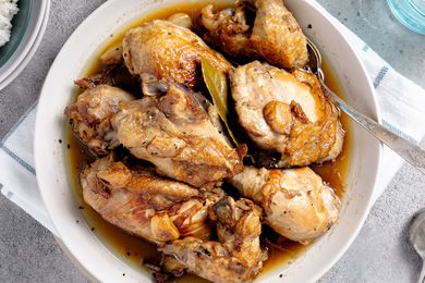 Chicken Adobo in a Bowl With a Spoon, Surrounded by a Table Setting With a Bowl of Rice, a Glass of Water, and More Utensils on the Counter