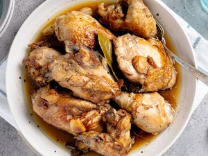 Chicken Adobo in a Bowl With a Spoon, Surrounded by a Table Setting With a Bowl of Rice, a Glass of Water, and More Utensils on the Counter