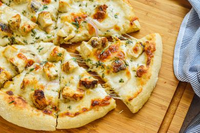 Chicken Alfredo Pizza Cut Into Slices With One Slice Slightly Removed From the Rest to Get a Cheesy Pool, All on a Cutting Board, and Next to the Board, a Blue and White Striped Kitchen Towel