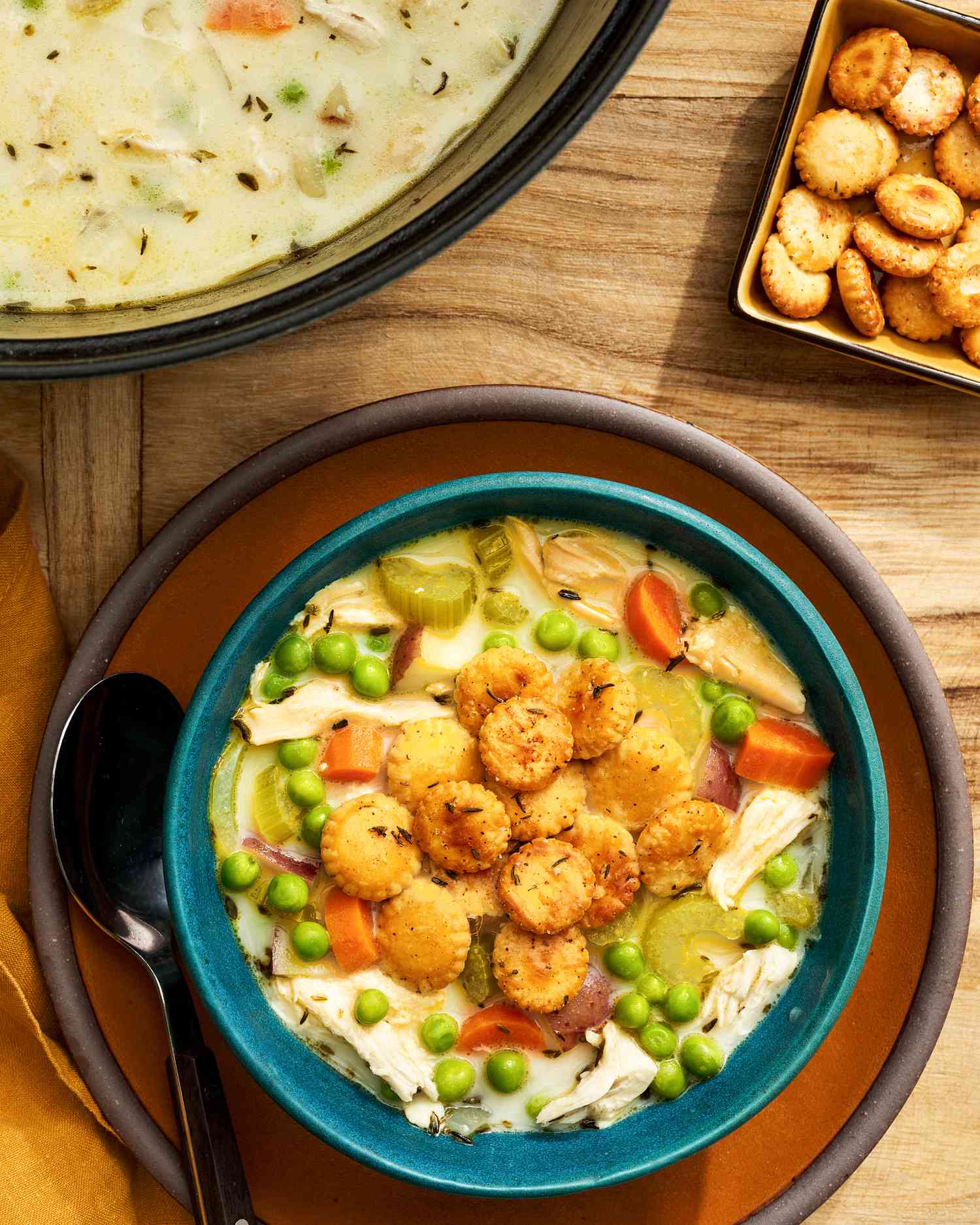 Bowl of chicken pot pie soup topped with toasted oyster crackers on a plate with a spoon and next to it, a pot with more chicken pot pie soup, a small bowl with more toasted oyster crackers, and mustard colored table napkin