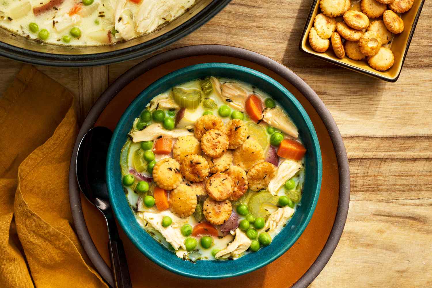 Bowl of chicken pot pie soup topped with toasted oyster crackers on a plate with a spoon and next to it, a pot with more chicken pot pie soup, a small bowl with more toasted oyster crackers, and mustard colored table napkin