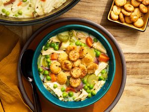 Bowl of chicken pot pie soup topped with toasted oyster crackers on a plate with a spoon and next to it, a pot with more chicken pot pie soup, a small bowl with more toasted oyster crackers, and mustard colored table napkin