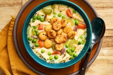 Bowl of chicken pot pie soup topped with toasted oyster crackers on a plate with a spoon and next to it, a mustard colored napkin