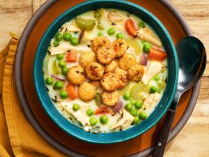 Bowl of chicken pot pie soup topped with toasted oyster crackers on a plate with a spoon and next to it, a mustard colored napkin