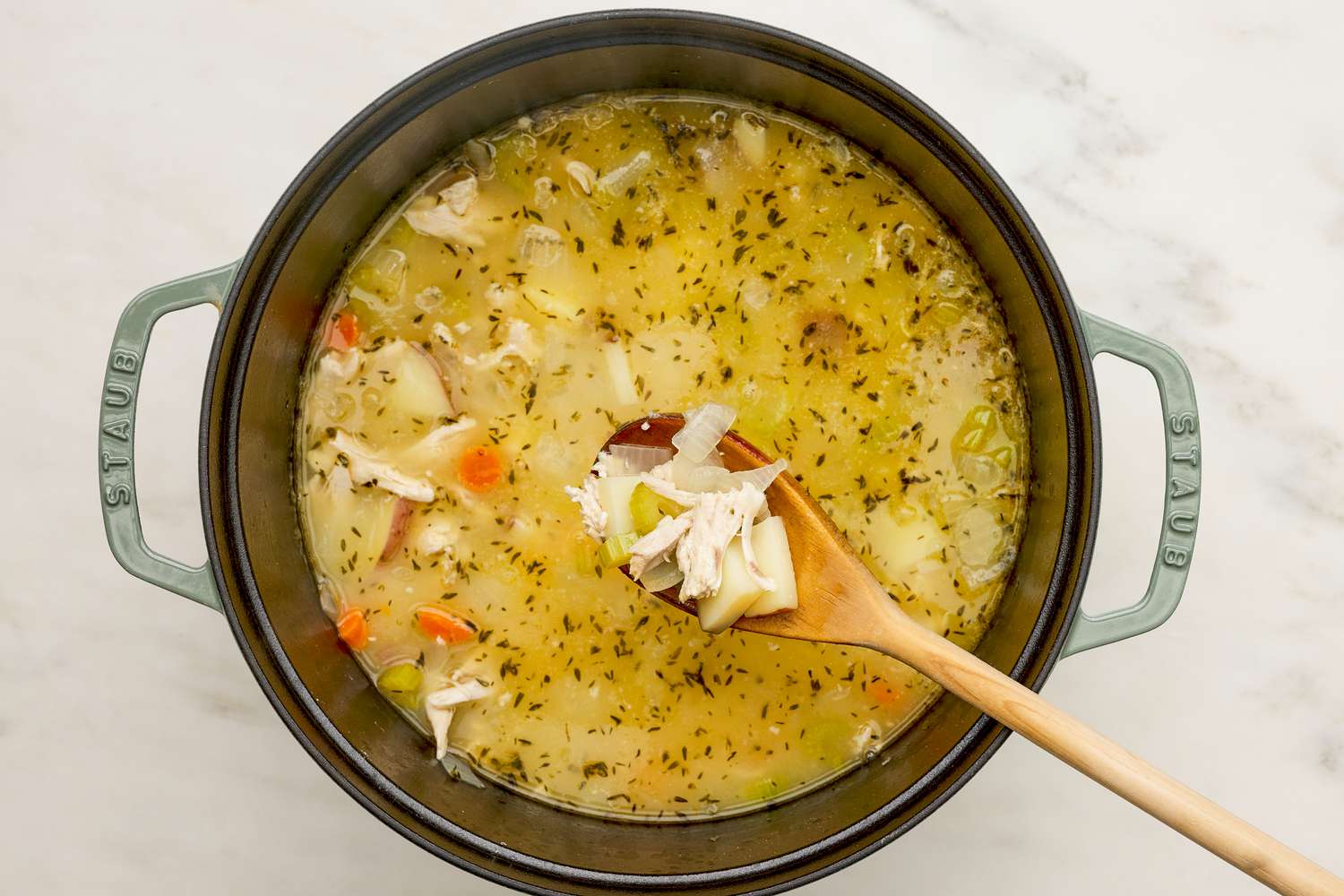 Seasoning, shredded chicken, and chopped potatoes, and chicken stock mixed into the vegetables in the Dutch oven and some lifted on the wooden spoon for chicken pot pie soup recipe