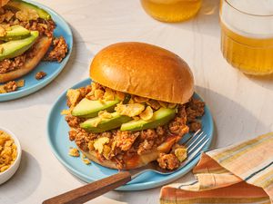 Chorizo sloppy joe (with avocado slices and crushed Fritos) on a plate with a fork at a table setting with another serving on another plate, two glasses of beer, a bowl of crushed Fritos, and a colorful stripped kitchen towel