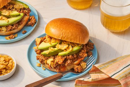 Chorizo sloppy joe (with avocado slices and crushed Fritos) on a plate with a fork at a table setting with another serving on another plate, two glasses of beer, a bowl of crushed Fritos, and a colorful stripped kitchen towel