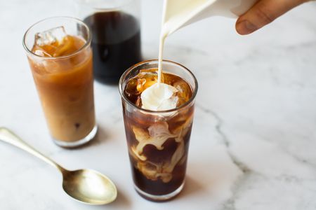 A hand pouring a pitcher of creamer into a glass of iced coffee
