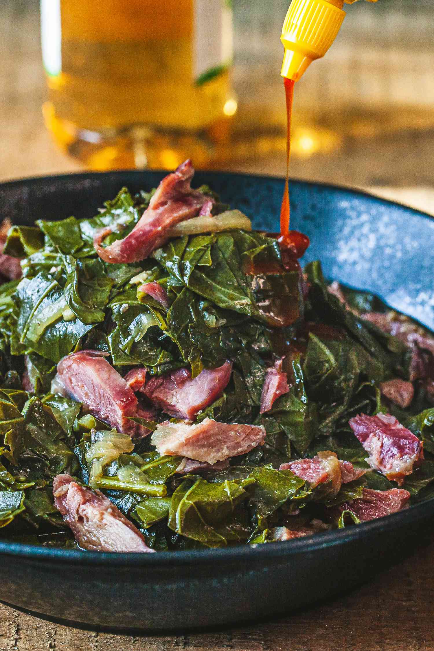 A big bowl of collard greens with pieces of ham hock. Hot sauce is being squeezed onto the top of the pile of collards.