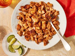 Bowl of copycat Chipotle chicken with a spoon, and in the surroundings, a bowl of lime quarters, glasses of water, and a scarlet table napkin