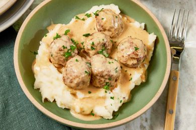 Bowl of copycat Ikea Swedish meatballs served over mashed potatoes, and in the surroundings, a table setting with a stack of plates and a sage table napkin